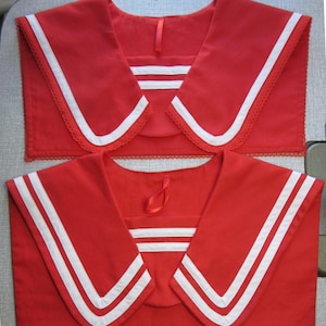 May include: Two red sailor collars with white stripes and trim. The collars have a classic design, with a square neckline and a wide, pointed collar. The top collar has a lace trim along the edge.