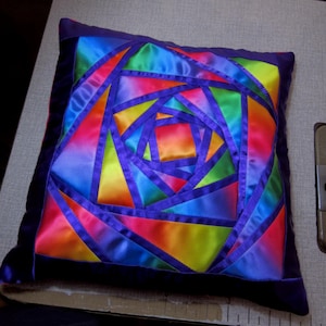 May include: A colourful pillow with a geometric design made of purple, blue, green, yellow, orange, and red satin fabric. The pillow is made of multiple pieces of fabric sewn together to create a spiral pattern.