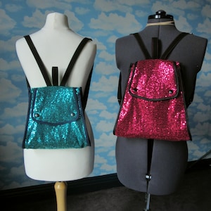 Puede incluir: Dos mochilas de lentejuelas, una turquesa y otra rosa, en maniquíes. Las mochilas tienen correas negras y un pequeño bolsillo en la parte delantera.