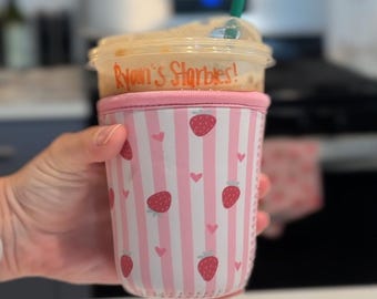 Könnte beinhalten: Ein kaltes Getränk in einem durchsichtigen Plastikbecher mit einem grünen Strohhalm. Der Becher befindet sich in einer rosa-weiß gestreiften Hülle mit Erdbeer- und Herzmotiven. Der Text "Ryan's Starbies!" ist auf dem Becher aufgedruckt.