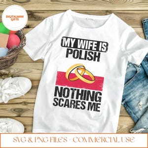 Könnte beinhalten: Weißes T-Shirt mit dem schwarzen Text "MY WIFE IS POLISH NOTHING SCARES ME". Das Design umfasst zwei goldene Eheringe über einem rot-weißen Streifen. Das T-Shirt liegt auf einer Holzoberfläche.