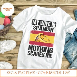 Op de afbeelding: Wit T-shirt met de tekst "MY WIFE IS SPANISH NOTHING SCARES ME" in het zwart. Het ontwerp bevat de Spaanse vlag en twee gouden trouwringen. Het shirt ligt op een houten oppervlak.