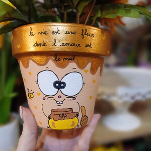 Peut inclure: Pot de fleur en terre cuite peint à la main avec un dessin d'abeille de dessin animé. Le pot est beige avec un bord doré et porte l'inscription française "la vie est une fleur dont l'amour est le miel". Il contient une plante.