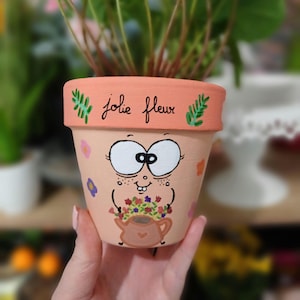 Peut inclure: Pot de fleurs en terre cuite peint à la main avec un dessin de visage de dessin animé. Le pot est de couleur pêche avec un bord orange clair et porte les mots "jolie fleur". Le visage de dessin animé a de grands yeux, des taches de rousseur et tient un bouquet de fleurs.