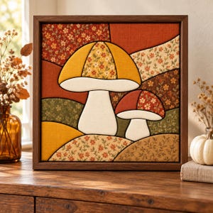 May include: A square framed fabric wall art piece featuring a patchwork design of two mushrooms. The mushrooms are constructed from various fabric segments in shades of mustard yellow, burnt orange, olive green, and cream with floral patterns.