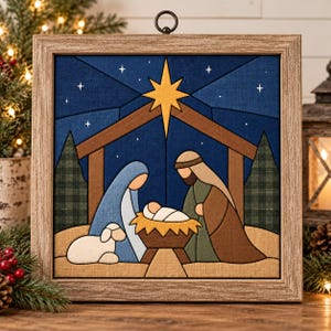 May include: A square framed fabric wall art piece depicting a nativity scene with a yellow star, a blue night sky, a brown stable, and figures in blue, green, and brown clothing. A small white lamb sits beside the figures near a manger.
