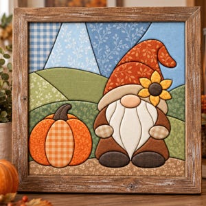 May include: A square fabric wall art piece in a rustic wooden frame featuring a quilted-style scene of a gnome with a sunflower on its orange hat and a large orange pumpkin. The background displays blue and green fabric patches with leaf patterns.