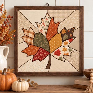 May include: A square wall art piece in a dark wood frame featuring a fabric quilt-style maple leaf design. The leaf is composed of various patterned fabric segments in shades of orange, red, green, brown, and cream, set against a light background.