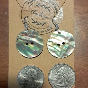 May include: Button-shaped earrings with iridescent shells, displayed on a brown card with the text "Thank you, Made with Love." Two quarters are shown below the earrings. The earrings are approximately 1.25 inches in diameter.