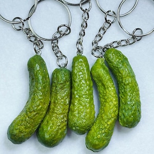Puede incluir: Cinco llaveros de pepinillos verdes con cadenas y anillos plateados. Los pepinillos tienen una superficie texturizada y diferentes tonos de verde. Los llaveros están dispuestos en fila.