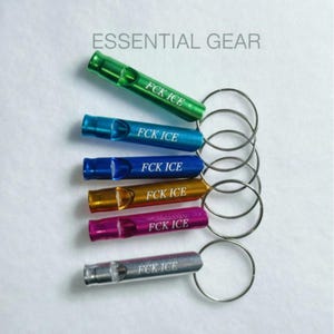 May include: An assortment of six metal whistles in various colors, including green, blue, gold, purple, and silver, each attached to a silver keyring. The text "FCK ICE" is printed on each whistle. The words "ESSENTIAL GEAR" are printed above the whistles.