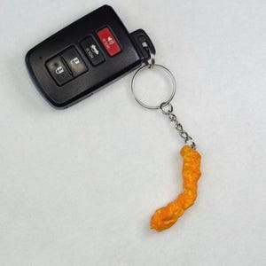 May include: Black car key fob with a silver keyring attached to a bright orange, curved, Cheeto-shaped keychain. The key fob has buttons for lock, unlock, and panic. The background is a plain, light-colored surface.