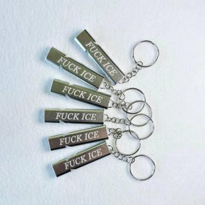 Puede incluir: Seis llaveros plateados con la inscripción "FUCK ICE". Los llaveros rectangulares con bordes redondeados están sujetos a una cadena y un anillo de metal. Están dispuestos sobre una superficie blanca.