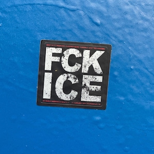 Op de afbeelding: Een vierkante sticker met de woorden "FCK ICE" in vette, witte letters op een zwarte achtergrond. De sticker heeft een rode rand en is geplaatst op een blauw oppervlak.