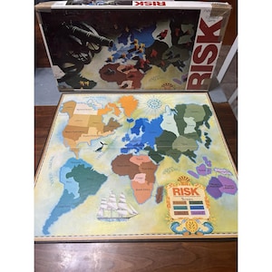 May include: A vintage Risk board game with a world map design. The game board features continents in various colors, with the game box visible above. The box has the word "RISK" in large red letters.