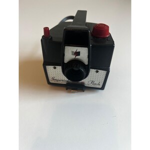 Könnte beinhalten: Eine Vintage-Schwarz Imperial Flash Kamera mit rotem Auslöser und Blitzkappe. Die Kamera hat einen Griff und die Wörter "Imperial" und "Flash" sind auf der Vorderseite aufgedruckt. Hergestellt in den USA.