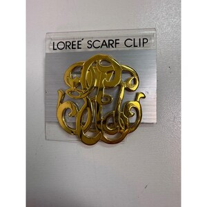 May include: A gold-colored scarf clip with an ornate monogram design. The clip is labeled "LOREE SCARF CLIP" in black text on a clear plastic backing. The monogram features intertwined letters with a glossy finish.