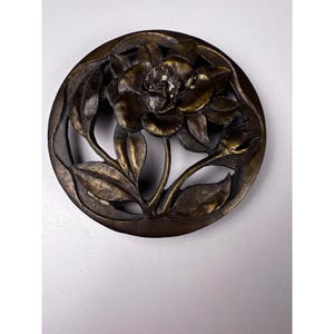 May include: A bronze-toned, circular decorative piece featuring a detailed floral design. The central flower is surrounded by leaves and stems, all within a circular frame. The piece has an antique or vintage aesthetic.
