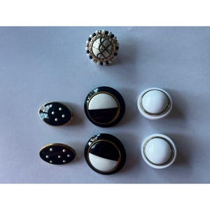 May include: A collection of decorative buttons in various shapes and designs. The buttons feature a mix of black, white, and gold colors. Some buttons have a glossy finish, while others have a textured appearance. The buttons are suitable for sewing or craft projects.
