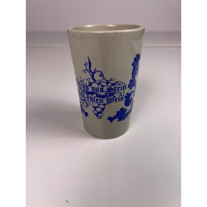 Puede incluir: Un vaso de cerámica gris con diseños azules de uvas y vides. El vaso tiene el texto azul "Trink aus Stein, edlen Wein". El vaso es cilíndrico y tiene una parte superior ligeramente ensanchada.