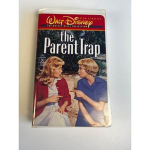 Puede incluir: Una cinta de VHS de la película de Disney "The Parent Trap". La portada presenta una pancarta roja y blanca con el logotipo de Walt Disney. El título está en letras blancas grandes. Dos mujeres están en la portada, señalándose.
