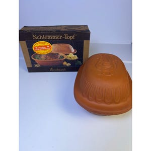 Puede incluir: Una olla de barro marrón Schlemmer-Topf con su tapa a juego, junto a su caja de cartón original. La caja presenta el nombre del producto y una imagen de comida cocinada. La olla tiene un diseño de gallo en la tapa.
