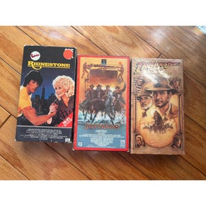 Puede incluir: Tres cintas de vídeo VHS de películas. Las cintas son para las películas "Rhinestone", "Silverado" e "Indiana Jones". La cinta "Rhinestone" tiene un fondo negro con el título de la película en amarillo. La cinta "Silverado" tiene un fondo rojo. La cinta "Indiana Jones" está sellada.
