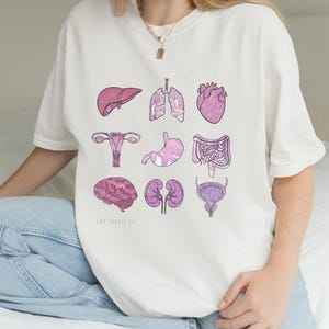May include: White t-shirt featuring a pink and purple illustration of human organs, including the liver, lungs, heart, uterus, stomach, intestines, brain, kidneys, and bladder. The shirt has short sleeves and a crew neck. The brand name "LAB ICONIC CO." is printed at the bottom.