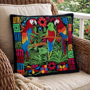 Puede incluir: Cojín cuadrado bordado sobre fondo negro con loros coloridos, flores y follaje. Los loros son rojos, azules y verdes. El cojín tiene un borde negro con un diseño geométrico en colores brillantes.