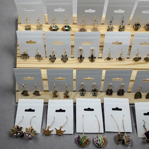 May include: A wooden display rack holds multiple pairs of Mardi Gras-themed earrings. The earrings feature various designs, including crowns, fleur-de-lis, and masks, in gold, green, purple, and yellow. Each pair is displayed on a white card with a hanging hole.