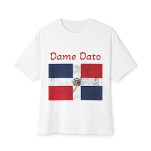 May include: White t-shirt featuring the Dominican Republic flag design. The flag is divided into four sections: two blue and two red, with a white cross and a coat of arms in the center. The text "Dame Dato" is printed above the flag.