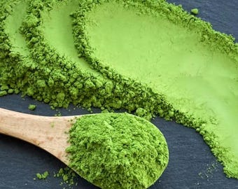 May include: Close-up of vibrant green matcha powder, with a wooden spoon filled with the powder. The matcha is arranged in a decorative pattern on a dark surface, showcasing its fine texture and vivid color.