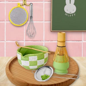 May include: A wooden tray displays a green and white chequered bowl, a stainless steel sieve with green powder, and a bamboo whisk. A green and bamboo matcha whisk and a wooden scoop are also present. A yellow sponge and a whisk hang on the wall.