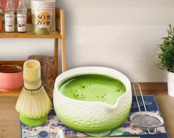 Può includere: Una ciotola per matcha in ceramica bianca e verde con beccuccio, riempita di tè verde. Accanto si trovano un frustino in bambù su un supporto verde, un cucchiaio in bambù su un poggia-cucchiaio bianco e un colino in metallo su un tappetino blu.