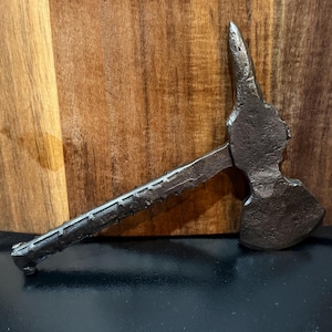 May include: A small, antique-style metal axe with a textured handle and a pointed end. The axe head has a curved blade and a spike on the opposite side. The axe is dark gray and rests against a wooden background.