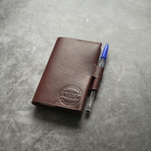 May include: A brown leather wallet with a pen holder and a blue pen. The wallet has a circular embossed logo on the front. The wallet is closed and rectangular.