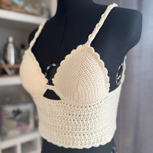 Könnte beinhalten: Cremefarbenes Häkeltop mit gewellten Kanten und geflochtenen Trägern. Das handgefertigte Kleidungsstück wird auf einer schwarzen Schaufensterpuppe präsentiert. Das Top hat ein strukturiertes Stichmuster und eine schmeichelhafte Silhouette.