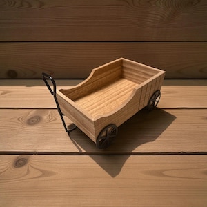 May include: A small, wooden wagon with a rectangular body and curved sides. The wagon has black metal wheels and a handle. The wood has a natural finish, and the wagon sits on a wooden surface.