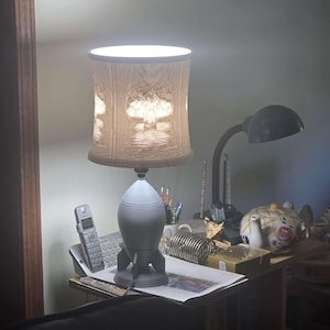 May include: A gray rocket-shaped lamp with a beige lampshade. The lampshade has a textured pattern. The lamp is on a desk with a telephone and other items. The lamp is turned on, emitting a bright light.
