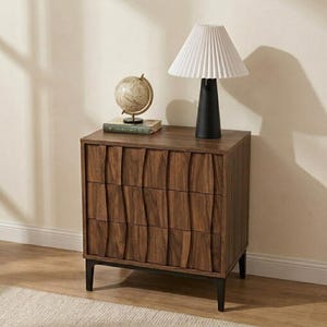 May include: A dark wood three-drawer chest with a textured, wavy front panel design and black legs. On top sits a table lamp with a black base and a white pleated conical shade, alongside a small globe resting on a book with a green cover.