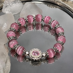 May include: A stretch bracelet featuring round, pink cat's eye beads alternating with ornate silver-toned metal spacers. The centerpiece is a circular silver-toned filigree charm set with a central oval pink gemstone surrounded by tiny white beads.