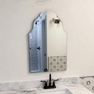 May include: A wall-mounted mirror with a decorative arched top and a smooth, frameless edge. The reflective surface shows a bathroom interior with gray tiled walls, a black faucet, and a white marble countertop with a patterned tile backsplash.