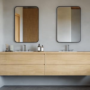 May include: A modern bathroom vanity with two rectangular mirrors with black frames. The light wood vanity has two sinks, two drawers, and two bottles of soap. A gray and white patterned rug and a pair of slippers are on the floor.