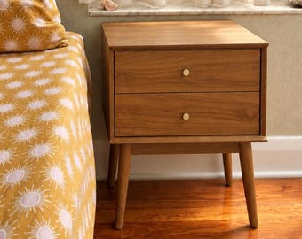 May include: A wooden nightstand with two drawers and small round gold knobs, standing on four tapered legs. Beside it is a bed covered in a mustard yellow fabric featuring a repeating pattern of white sunbursts and circular motifs.
