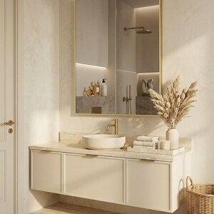 May include: A wall-mounted bathroom vanity with a cream-colored cabinet, a stone countertop, and a round vessel sink. Above the vanity is a rectangular mirror with a thin gold frame. A gold faucet, a tray with folded towels, and a vase with dried flowers.