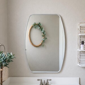 May include: A rectangular wall mirror with rounded corners and a thin silver frame. The mirror reflects a wooden oval wreath adorned with green faux succulents. The mirror is positioned above a white bathroom vanity sink with a metal faucet.