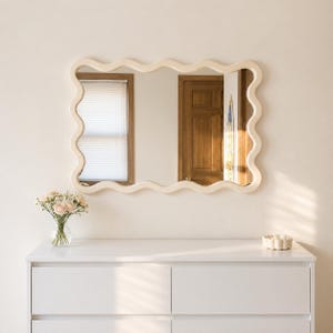 May include: A rectangular wall mirror with a wavy, cream-colored frame. The mirror reflects a room with a wooden door and a window with blinds. The mirror is mounted above a white dresser, which holds a glass vase with light pink flowers.