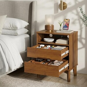 May include: A wooden nightstand with two open drawers and an open shelf. The top surface holds a cylindrical lamp, a smartphone, and a book. The drawers contain various small items including earbuds, cables, and folded fabric items.