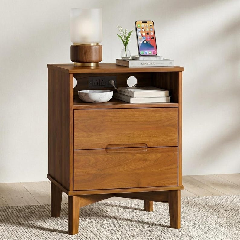May include: A wooden nightstand with two drawers and an open top shelf featuring a built-in power outlet. The nightstand holds a cylindrical lamp, a small vase with flowers, a smartphone, and two stacked books on its top surface.