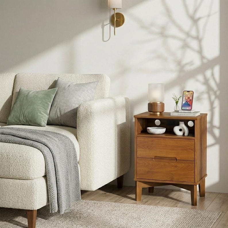 May include: A wooden nightstand with an open shelf and two drawers, featuring a cylindrical lamp, a small white ceramic sculpture, and a smartphone on top. Beside it sits a cream-colored textured sofa with a sage green pillow and a gray throw.